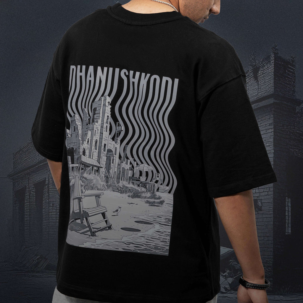 Person wearing Dhanushkodi black oversized t-shirt with a graphic design on the back, standing in front of a dark, urban background.