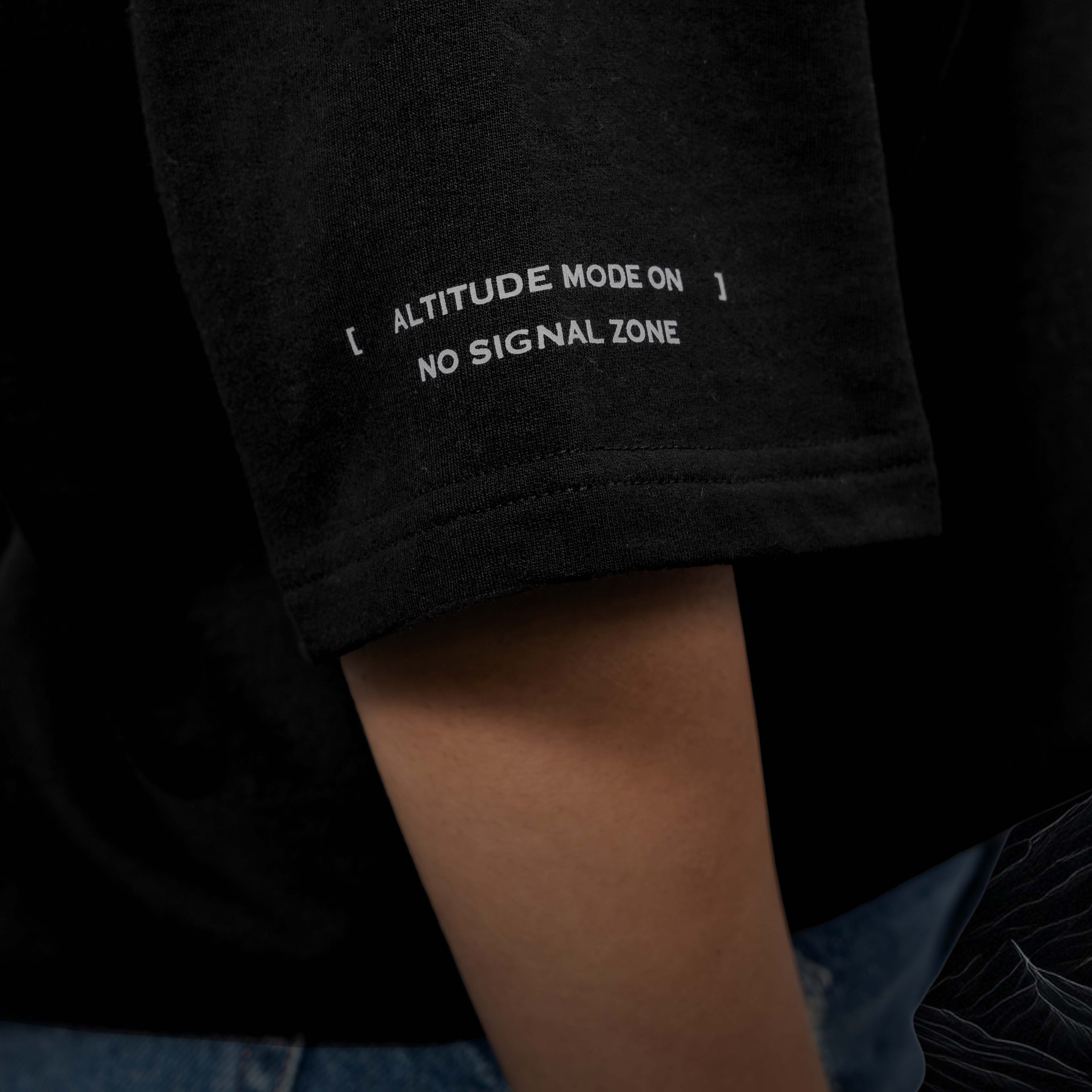 Kargil black Oversized t-shirt sleeve with white text 'ALTITUDE MODE ON NO SIGNAL ZONE' held by a person wearing jeans.
