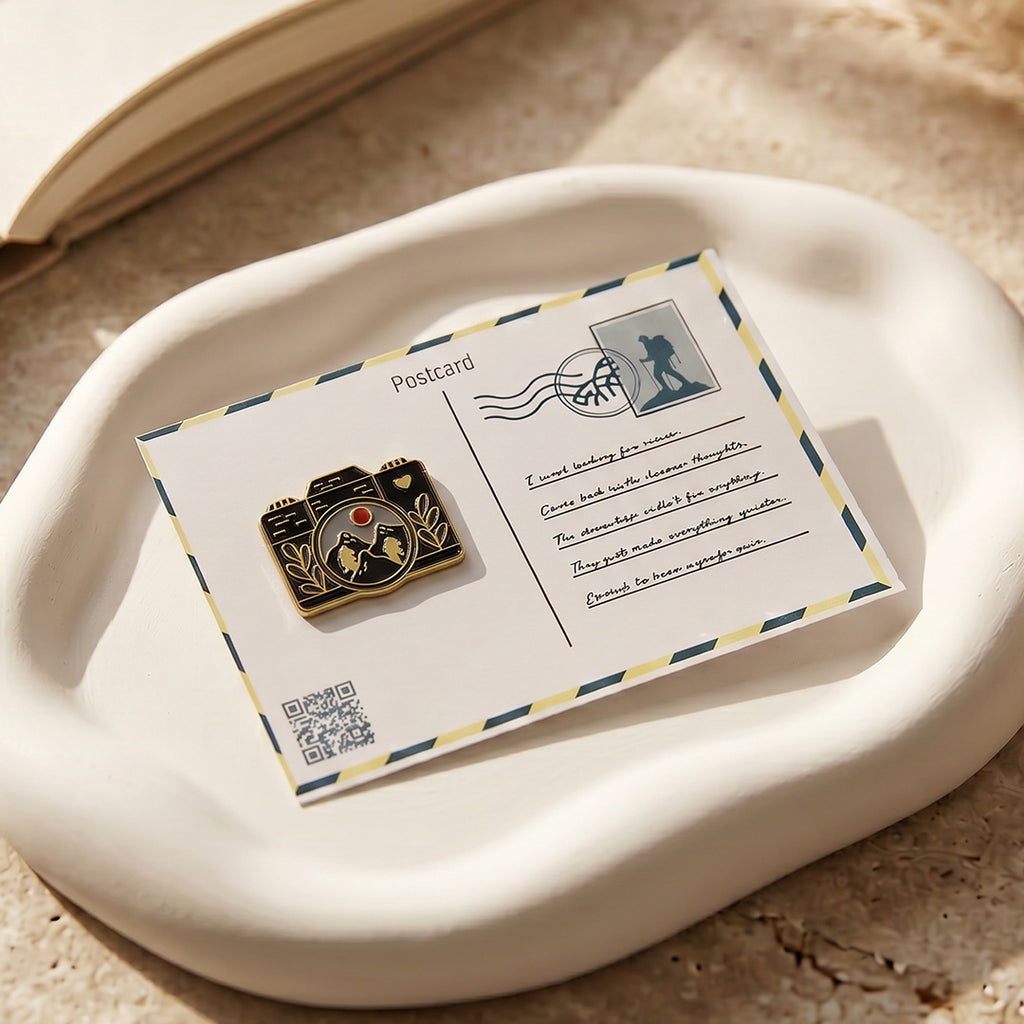 Camera-shaped pin on a postcard with a scenic illustration, placed on a textured surface.