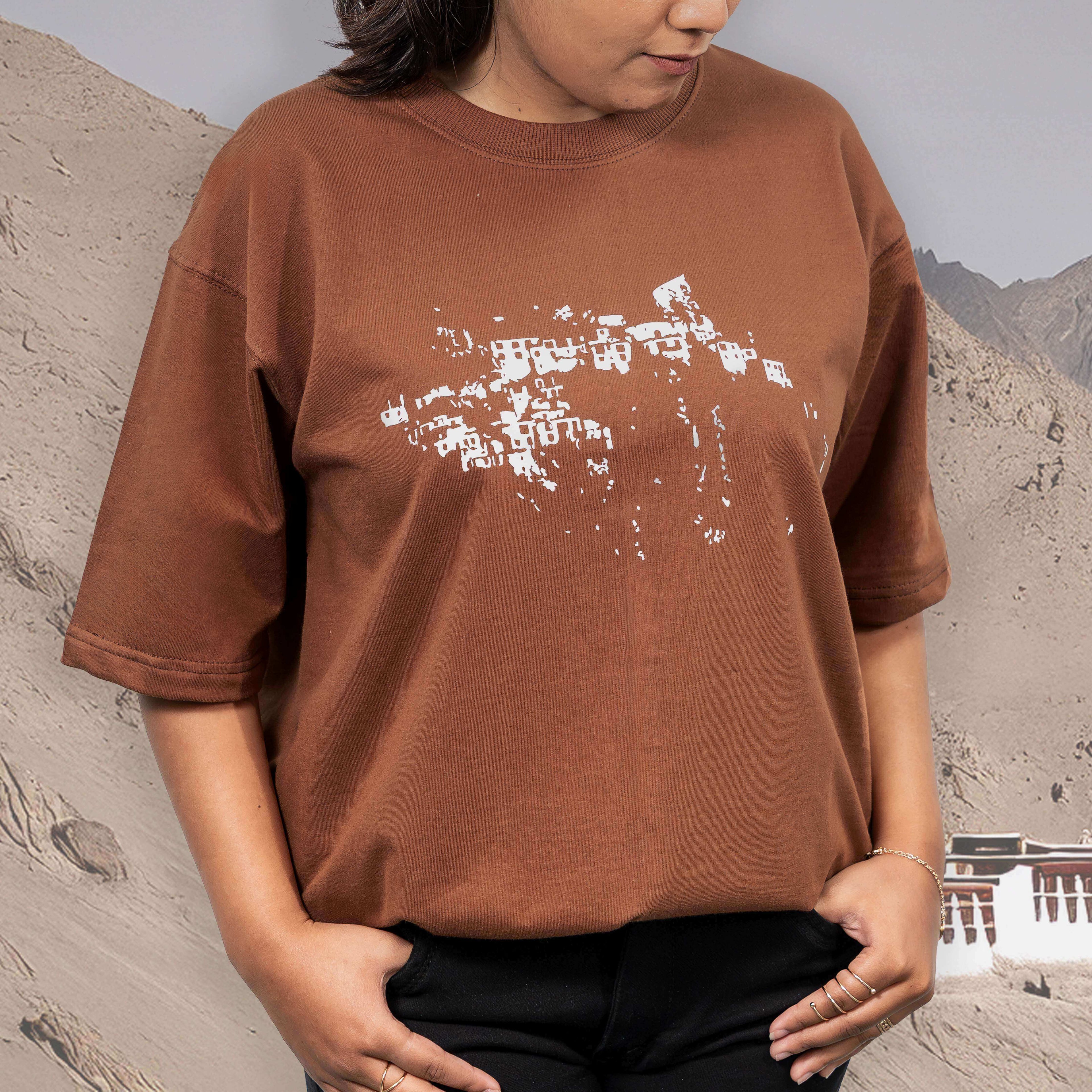 Person wearing a Leh brown oversized t-shirt with a white graphic design, standing against a mountainous background.