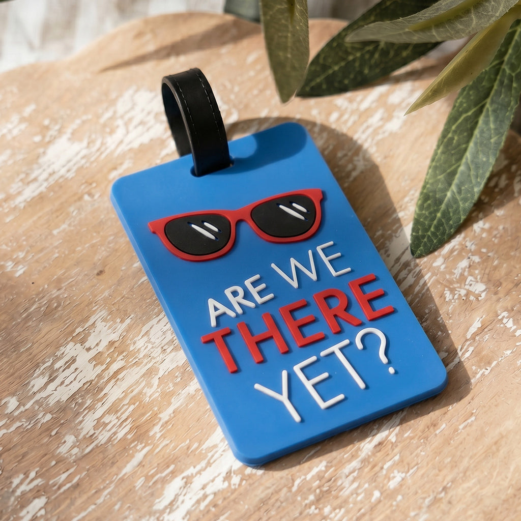 Blue luggage tag with sunglasses graphic and text 'ARE WE THERE YET?' on a wooden surface.