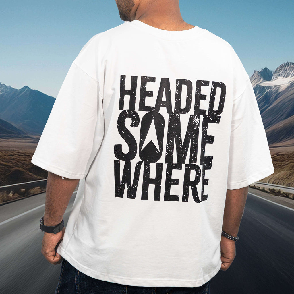 Person wearing a mountain helmet white oversized t-shirt with 'HEADED SOMEWHERE' text, standing on a road with mountains in the background.