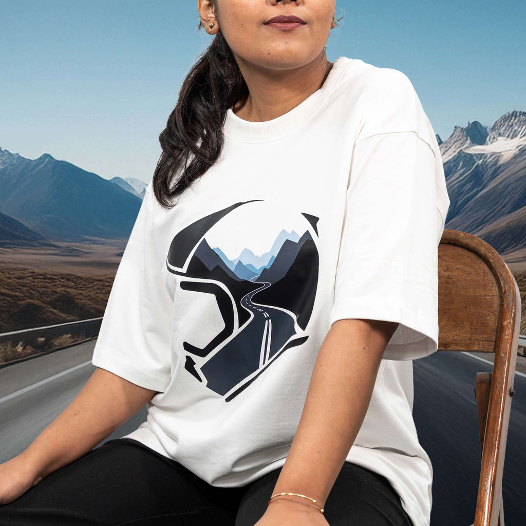 Person wearing a mountain helmet white oversized t-shirt with a graphic design of a helmet and mountain, sitting outdoors with mountains in the background.