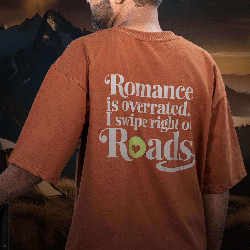 Person wearing Tinder for roads brown oversized t-shirt with 'Romance is overrated. I swipe right on roads' text in a scenic outdoor setting.