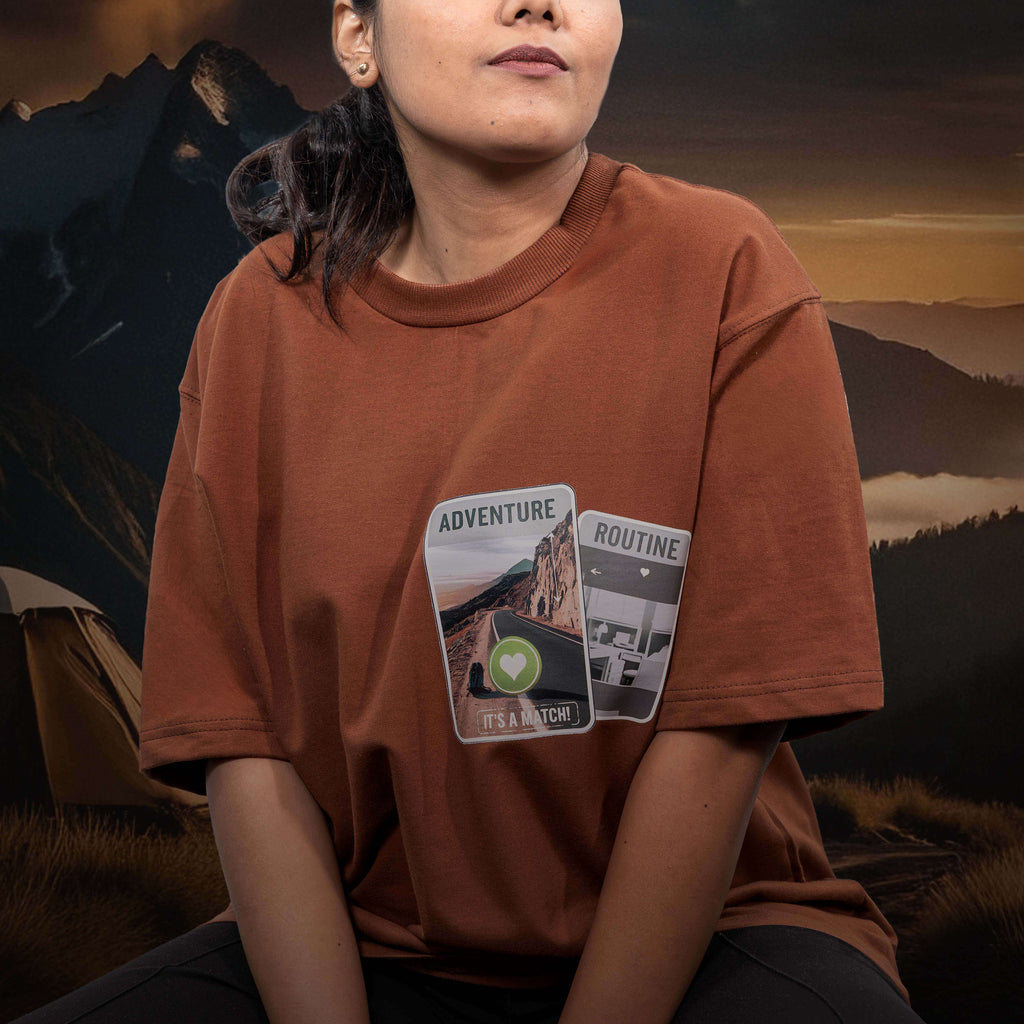 Person wearing tinder for roads brown oversized t-shirt with patches against a mountainous landscape background