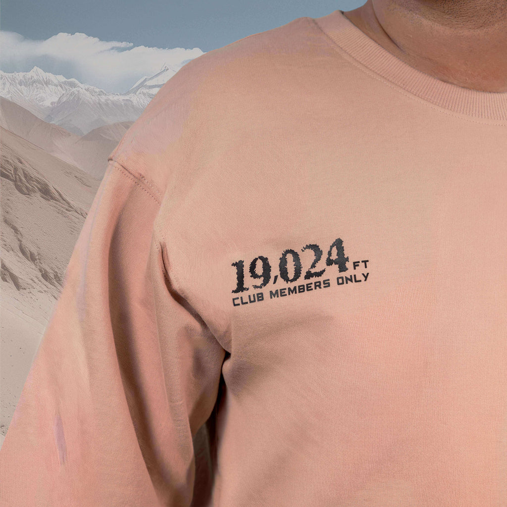 Person wearing an Umling La Beige Oversized t-shirt with '19,024 FT CLUB MEMBERS ONLY' text against a mountainous background