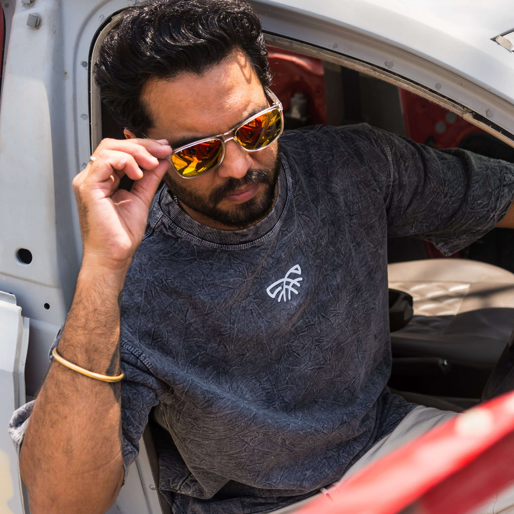 Man adjusting sunglasses inside a vehicle, wearing a acid wash t-shirt with a logo.