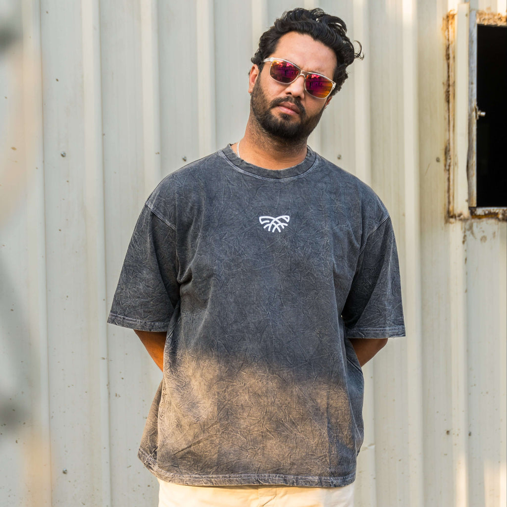 Man wearing acid wash t-shirt with a logo, standing against a corrugated metal wall.