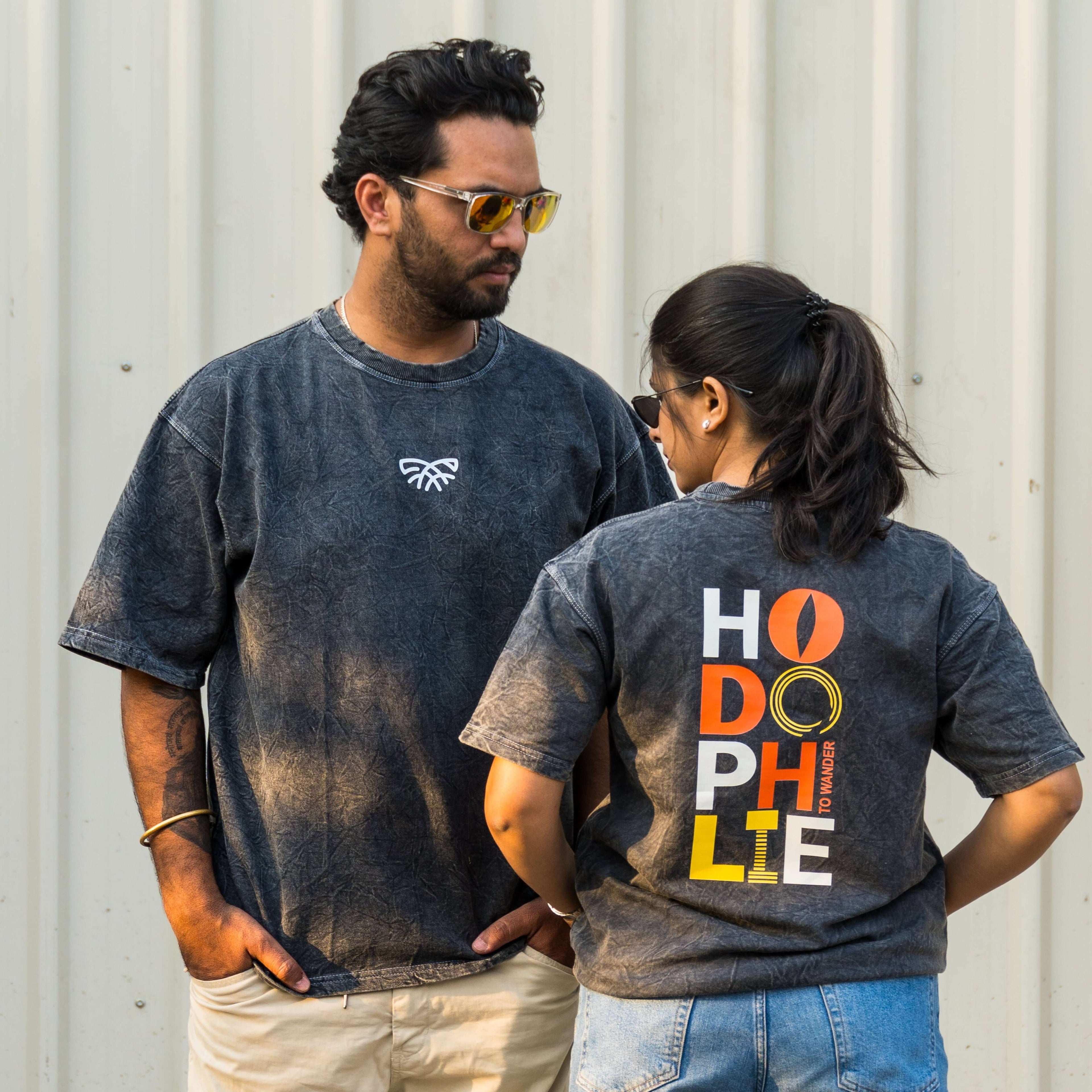Two people wearing acid wash t-shirt with hodophile text, standing against a corrugated metal wall.