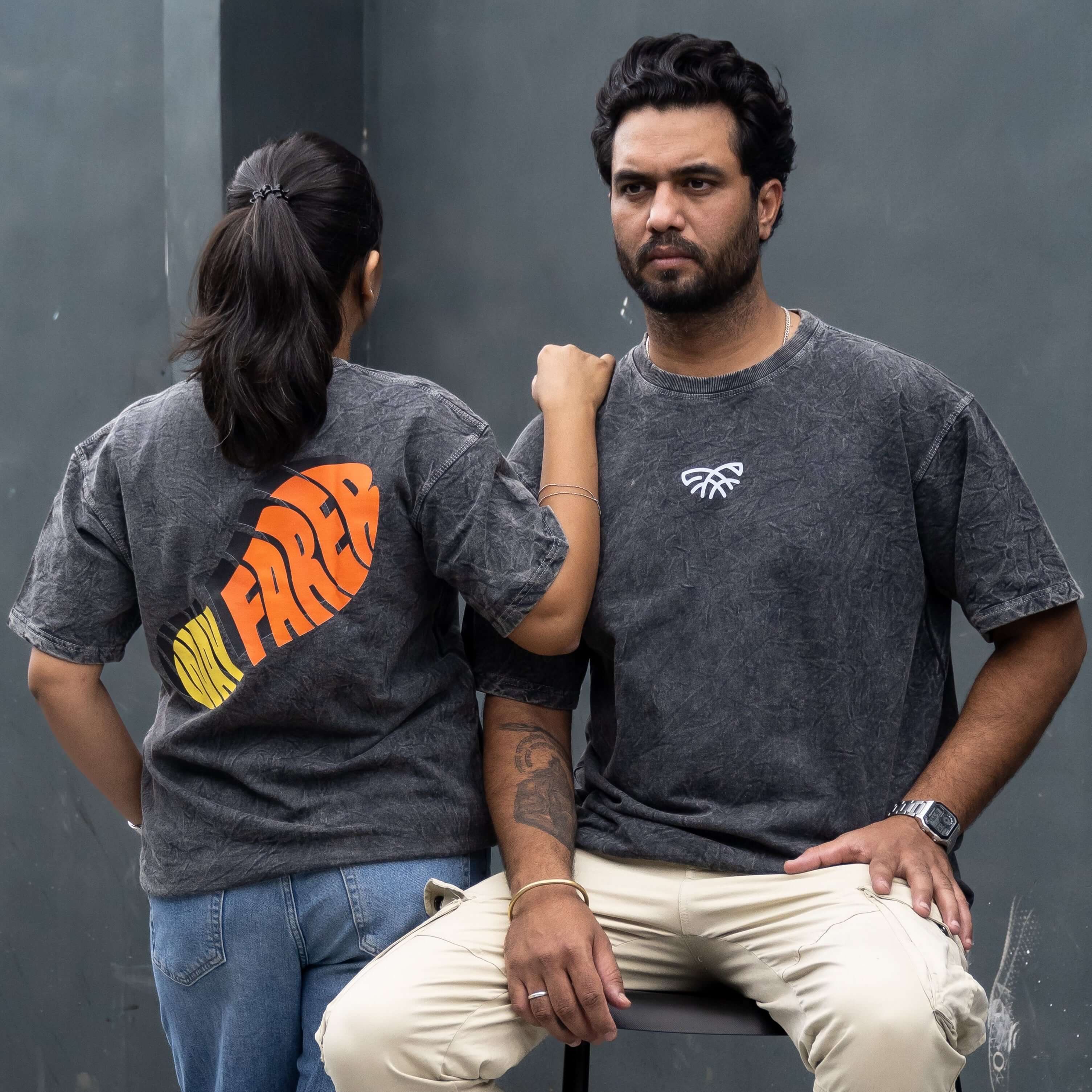 Two people standing and sitting on a stool against a dark wall wearing acid wash t-shirt showing front and back design
