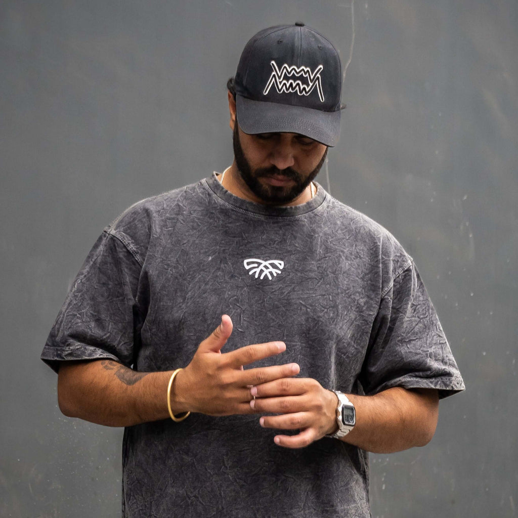 Man wearing acid wash t-shirt, cap, and beige pants against a gray wall.