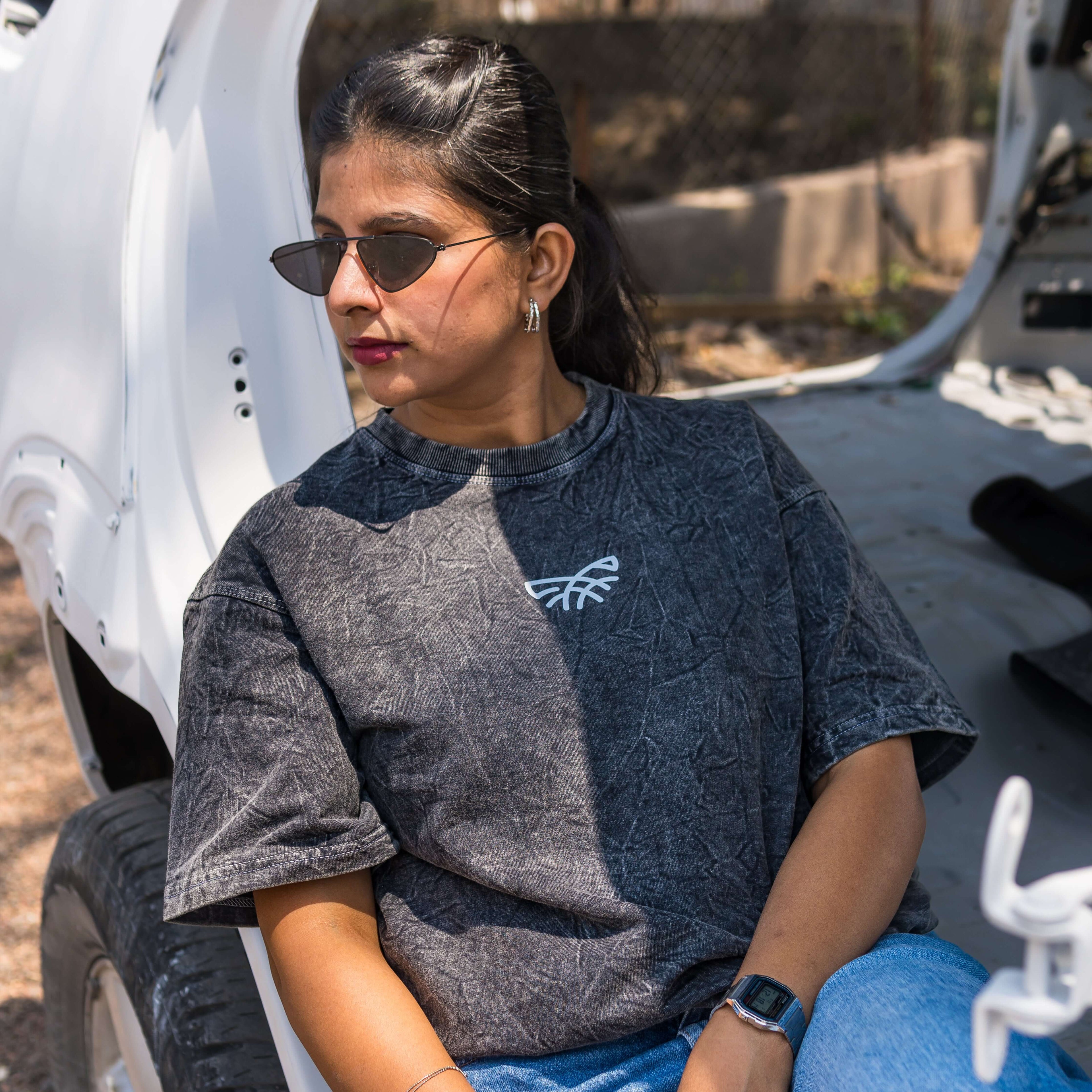 Woman wearing sunglasses and acid wash oversized t-shirt with a logo, sitting next to a white vehicle.